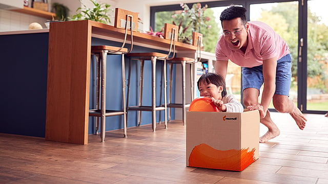 A child sitting in a cardboard box with Mondi branding, which he is pretending is a car and is being pushed by his dad.