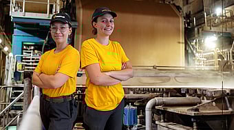 two female Mondi mill workers stand back to back with arms folded in a factory setting whilst wearing yellow t-shirts and black caps
