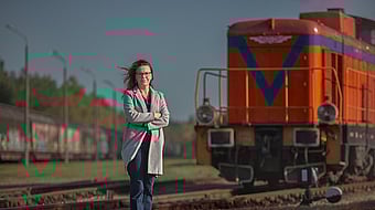 lady in grey coat and blue jeans with arms folded stands in front of a red train