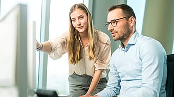 A man and a woman look closely at a computer screen; the woman points at it while both appear highly focused and engaged.