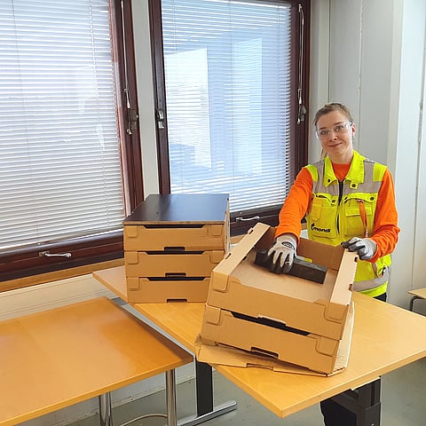A worker wearing high-visibility workwear and protective gloves sorts and folds cardboard packaging on a table in an office or warehouse workspace. Several cardboard boxes and packaging components are arranged on the work surface.