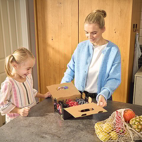 A woman and a child stand next to a kitchen island, opening a wrap-around container for fruits made of corrugated board.