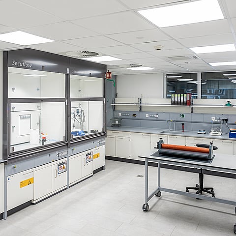 A clean laboratory workspace with workbenches, testing equipment and storage cabinets. A central worktable with a machine or testing device is positioned in the middle of the room, while large windows provide natural light.