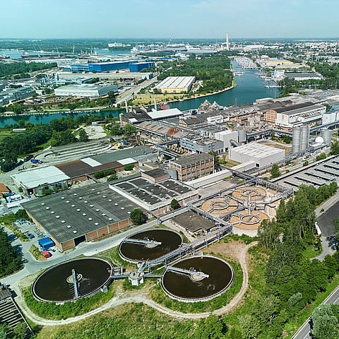 Aerial view of an industrial facility with large circular water-treatment basins, warehouses, and surrounding greenery near a river.