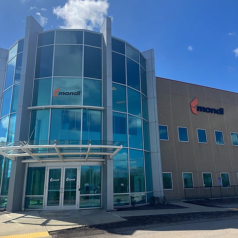 A modern office and industrial building with a glass-fronted entrance and multi-storey curved façade. The building features large windows, company signage and a paved exterior area under a clear blue sky.