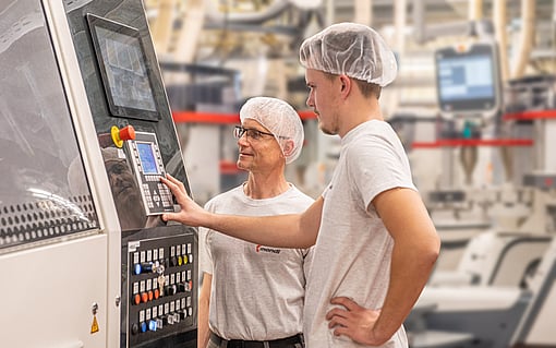 Two employees wearing protective hair coverings operate and monitor an industrial production machine in a manufacturing facility. One employee interacts with the control panel while the other observes the machine settings.