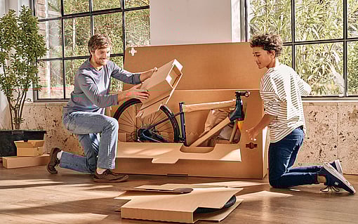 A man and a boy unboxing a bicycle from paper-based packaging.