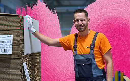 factory working looking into camera while wearing an orange shirt and blue overalls and leaning on a pallet of cardboard packaging