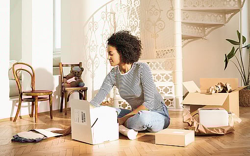 A woman sitting on the floor opening a corrugated box. Other corrugated boxes surround her, as well as a paper mailer bag and filling material made of paper.