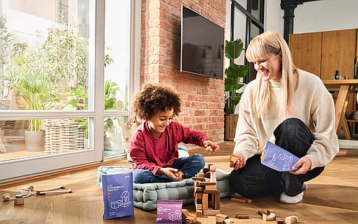 A young child and an adult sit on a wooden floor indoors, building a structure with wooden blocks. Cushions, wooden toys and several blue and purple activity boxes are placed around them. Large windows, indoor plants and a brick wall are visible in the background.