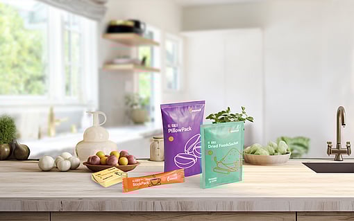 A clean, modern kitchen setting with soft natural light. On the wooden countertop, several Mondi flexible packaging samples are displayed upright, including a purple ‘PillowPack’, a green ‘Dried FoodSachet’, and an orange ‘StickPack’. Fresh vegetables and kitchen items, such as tomatoes, onions, lemons and a ceramic oil bottle, are arranged around the packs, creating a warm atmosphere.