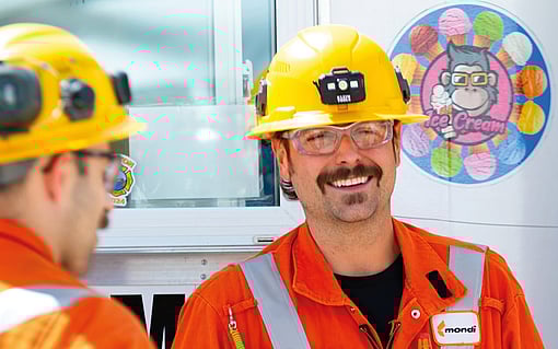 Two Mondi workers wearing orange safety uniforms and yellow hard hats stand together in an industrial setting, engaged in conversation near machinery.