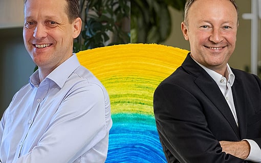 Two people standing with folded arms in front of a colourful circular display, with indoor plants and office elements visible in the background.