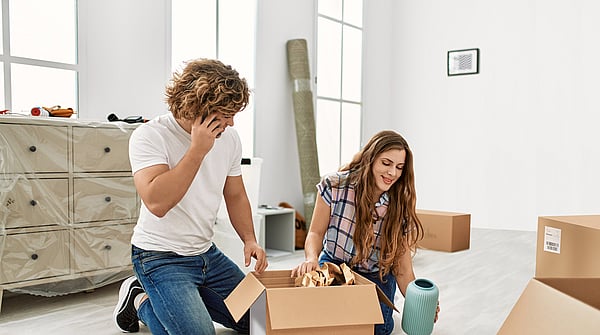 a man and a woman on their hands and knees unboxing a box in their home with the woman holding a small vase and the man on his mobile phone