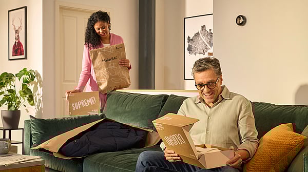 A man is sitting on the sofa unpacking a Mondi DelightBox with a Protective Mailer in front of him, while a woman stands in the back holding a MailerBag and a SupremeBox.