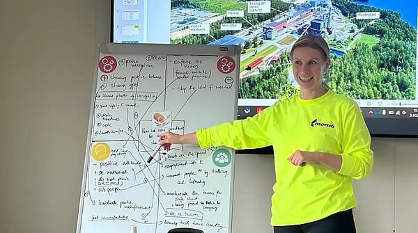 woman in bright jumper standing in front of a screen and a whiteboard whilst addressing an audience