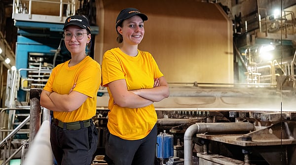 two female Mondi mill workers stand back to back with arms folded in a factory setting whilst wearing yellow t-shirts and black caps