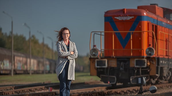 lady in grey coat and blue jeans with arms folded stands in front of a red train