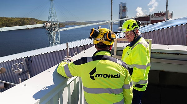 two men in hi-viz clothing overlook a production site with a river and forest in the background