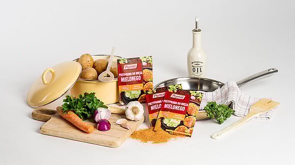 Assorted cooking ingredients arranged on a white background, including potatoes in a yellow pot, fresh vegetables like carrots, parsley, onions, and garlic on a wooden board, packets of Prymat seasoning for minced meat, a frying pan with a kitchen towel, and an olive‑oil bottle.
