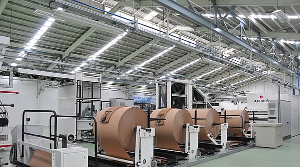 Industrial production line with large rolls of brown paper mounted on automated unwinding machines inside a factory hall.
