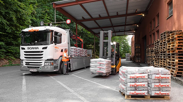 Delivery truck parked under a covered loading area while a forklift transports wrapped pallets. Stacks of palletised building materials are arranged in the foreground, with additional wooden pallets stored beside an industrial building surrounded by trees.