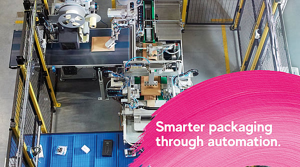 Overhead view of an automated packaging production line in an industrial facility, with machinery processing cardboard packages. A pink graphic overlay in the foreground displays the text: “Smarter packaging through automation.”