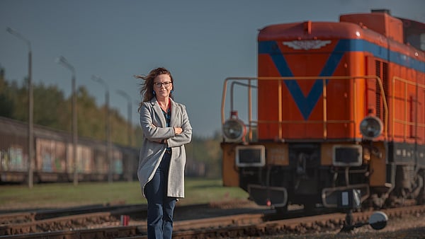 lady in grey coat and blue jeans with arms folded stands in front of a red train