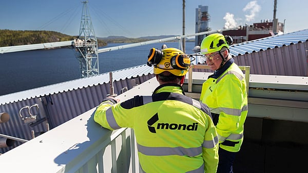two men in hi-viz clothing overlook a production site with a river and forest in the background