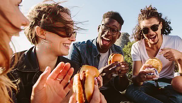 a group of young adults sitting in a group laughing, smiling and eating burgers