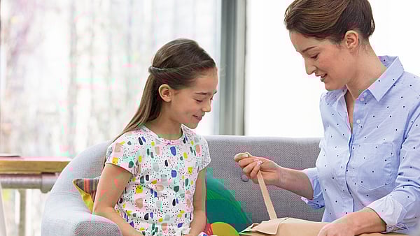 A child and an adult sit together on a sofa in a bright living room. The adult is opening a cardboard package, while the child watches attentively. Soft furnishings and natural light create a warm, home‑like atmosphere.