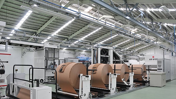 Industrial production line with large rolls of brown paper mounted on automated unwinding machines inside a factory hall.
