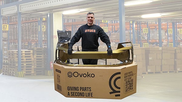 Person standing behind a large cardboard shipping box while holding a gold-coloured automotive bumper part placed across the top. The box is printed with the “Ovoko” logo and the text “Giving parts a second life,” along with a QR code and handling icons. The person is wearing dark clothing and gloves. Additional packaging material and a wrapped item are visible behind the box, all set against a plain white background.