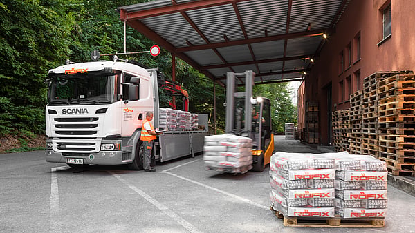 Delivery truck parked under a covered loading area while a forklift transports wrapped pallets. Stacks of palletised building materials are arranged in the foreground, with additional wooden pallets stored beside an industrial building surrounded by trees.