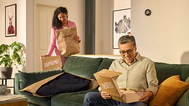 A man is sitting on the sofa unpacking a Mondi DelightBox with a Protective Mailer in front of him, while a woman stands in the back holding a MailerBag and a SupremeBox.