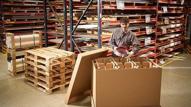 Mondi worker in a warehouse places a packaged item into a large cardboard box, surrounded by wooden pallets and metal shelving with stored materials.