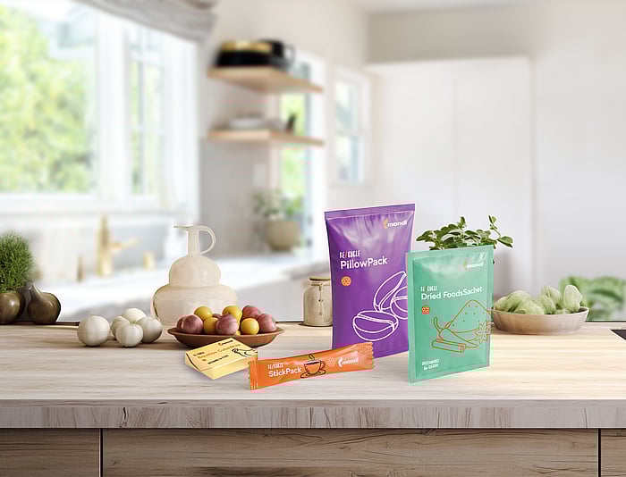 Auf einer holzfarbenen Küchenarbeitsplatte sind verschiedene farbenfrohe Verpackungsmuster ausgestellt, darunter ein lila Kissenbeutel, ein grüner Beutel für Trockenprodukte, ein orangefarbener Stickpack sowie zwei kleine, butterähnlich verpackte Artikel. Im Hintergrund stehen frisches Obst und eine Topfpflanze.