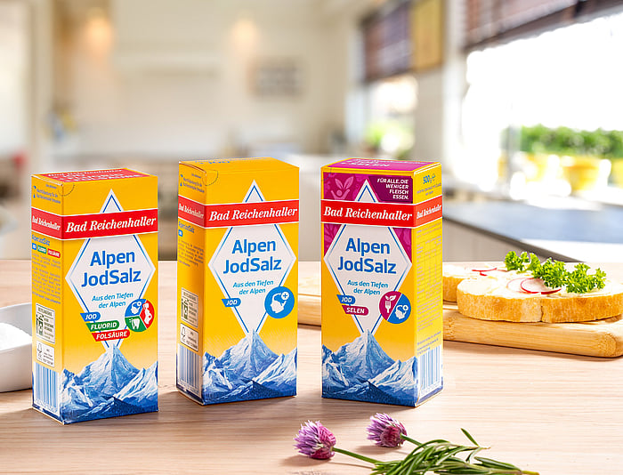 Drei Packungen „Alpen JodSalz“ stehen auf einer hölzernen Küchenarbeitsplatte. Die Verpackungen sind in leuchtenden Gelb-, Rot- und Blautönen gestaltet und zeigen eine Illustration alpiner Berglandschaften. Frische Kräuter und Küchenutensilien sind im Hintergrund weich verschwommen zu erkennen.