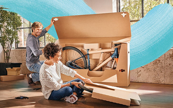 A man and a boy open a cardboard box containing a bike. A Mondi brush stroke graphic is imposed.