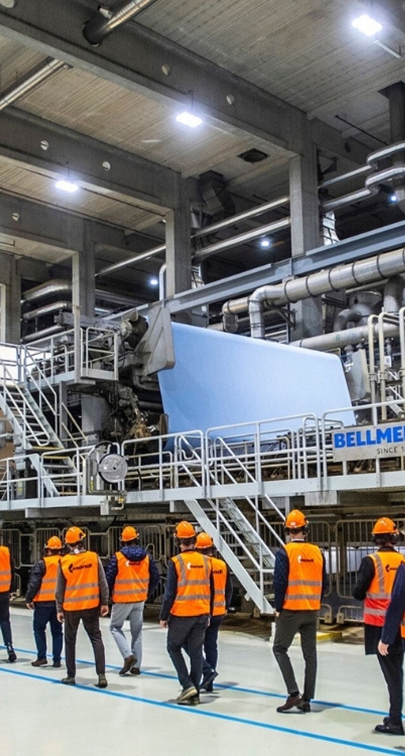 Employees wearing high-visibility vests and hard hats walk through a large industrial production hall with extensive machinery, pipes, and metal platforms.