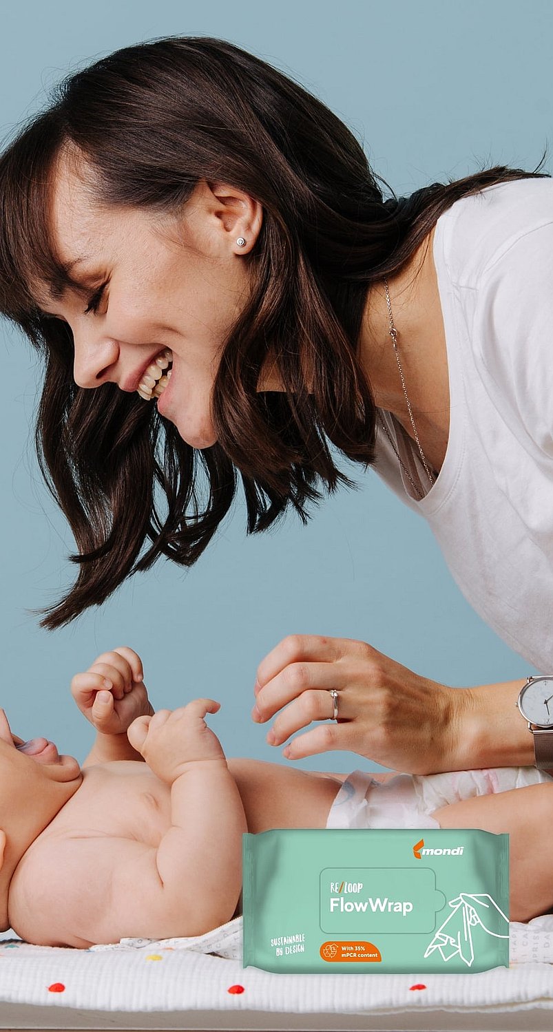 Eine Person beugt sich über ein Baby, das auf einer Wickelunterlage liegt. Im Vordergrund ist eine Packung Babyfeuchttücher zu sehen, links steht eine Babyflasche vor einem hellblauen Hintergrund.