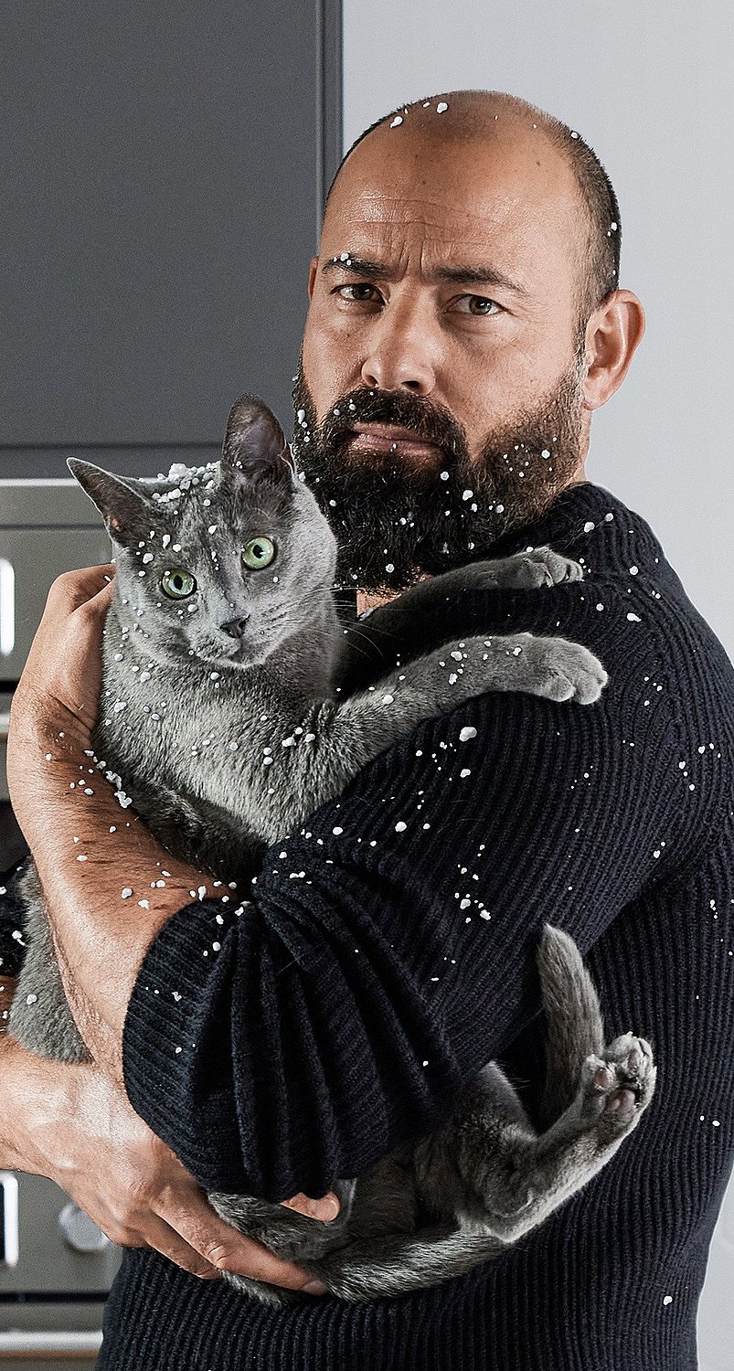 A man is standing in front of an oven, holding a cat. His beard and pullover are covered with pieces of polystyrene.