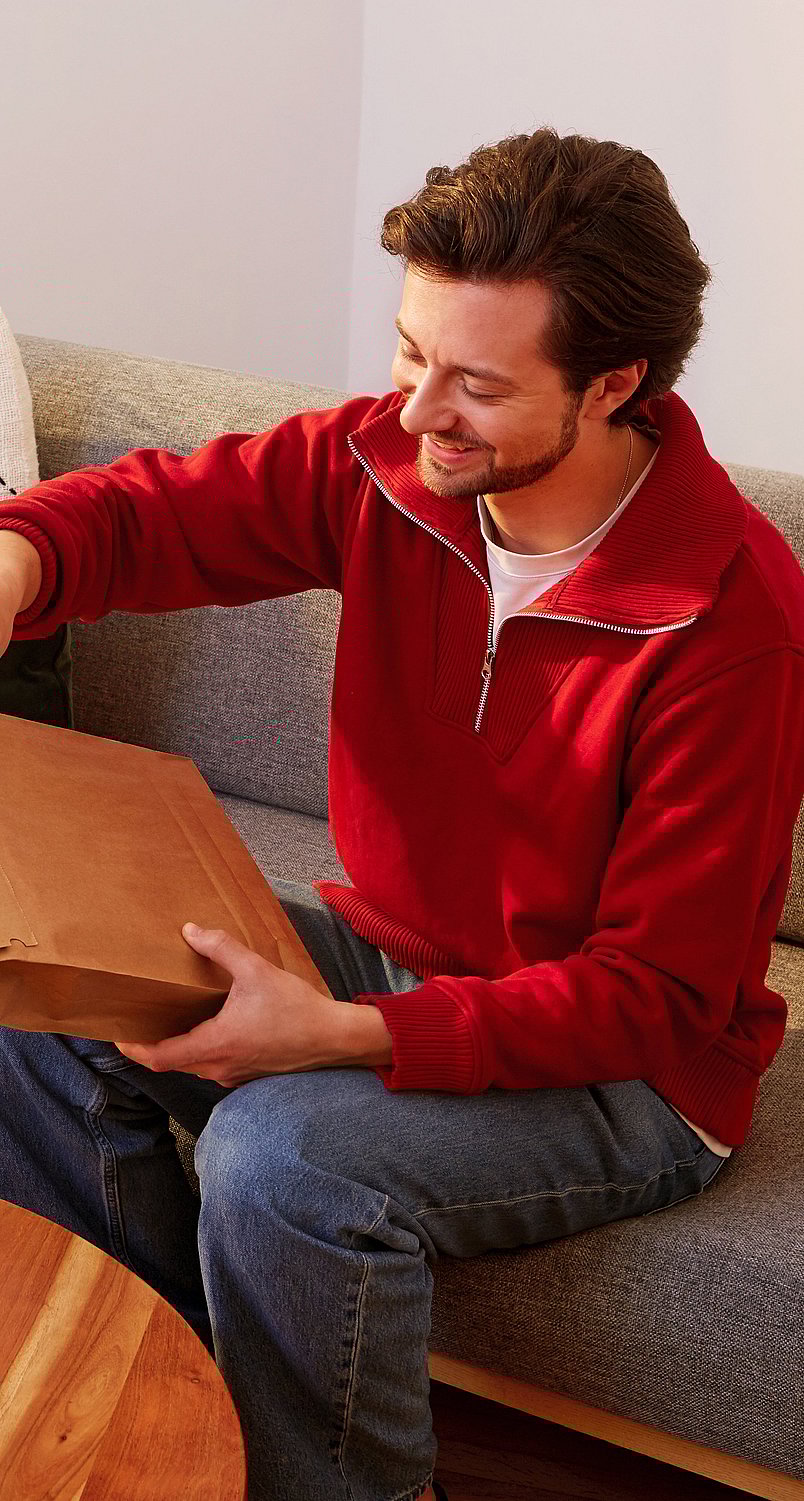 man in red jumper sitting on a green sofa opening a paper mailer package
