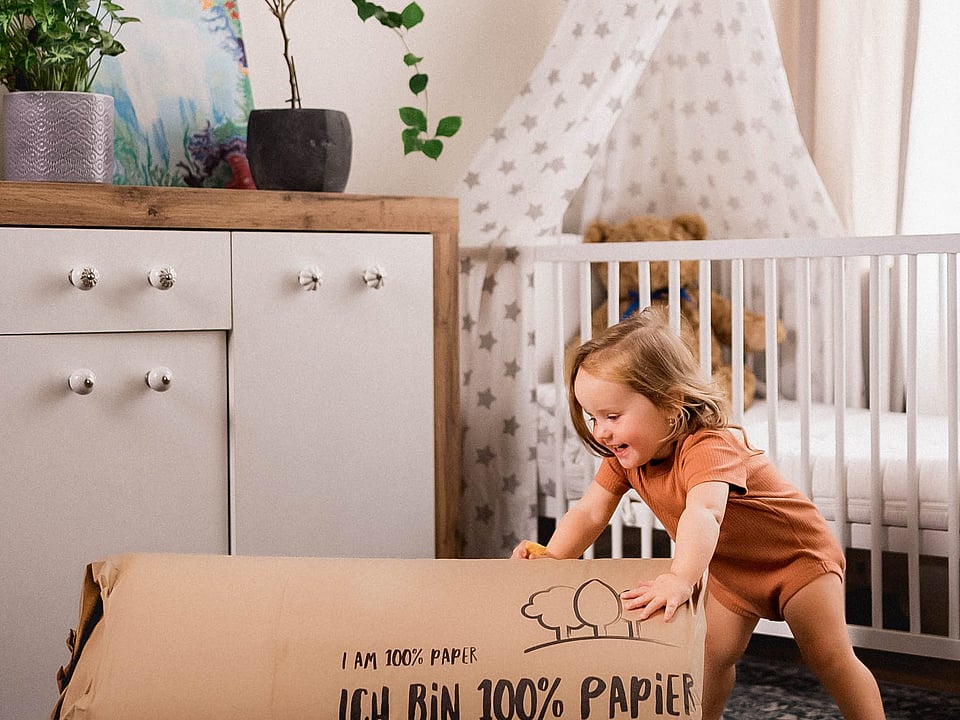 young girl in orange t-shirt and orange shorts leaning on a large piece of cardboard packaging and smiling with a crib behind her