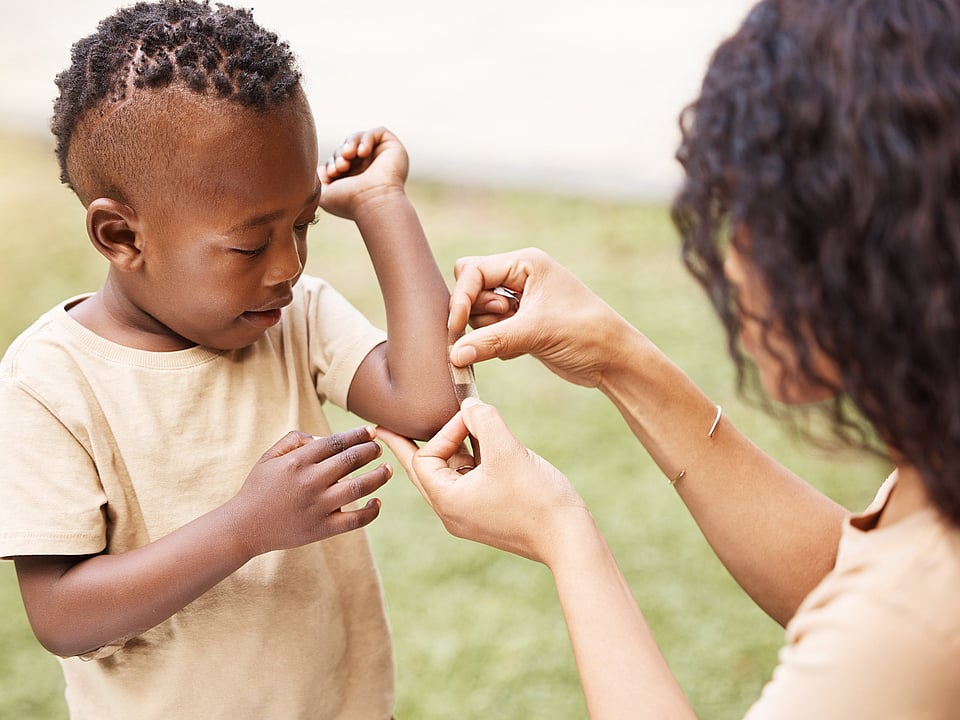 young child in beige t-shirt getting a plaster stuck on his left arm by a woman