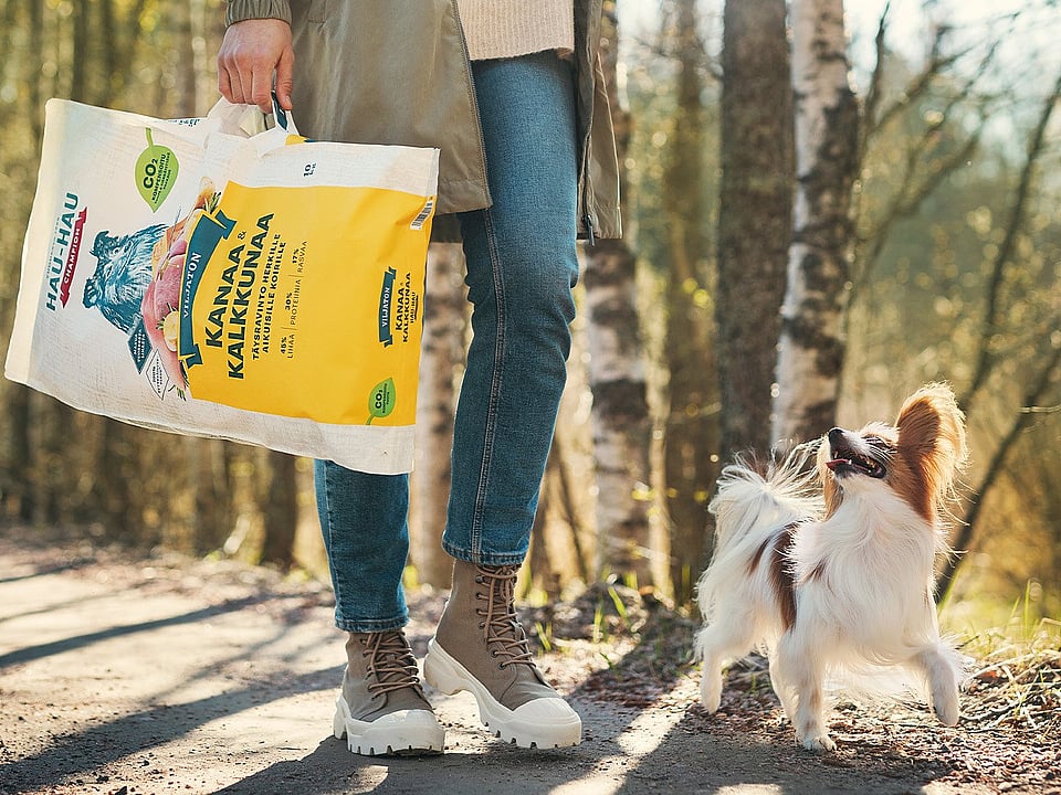 The bottom half of a lady wearing blue jeans, khaki jacket and brown and white boots walking beside a white and brown dog in a forest background while holding a bag of dog food