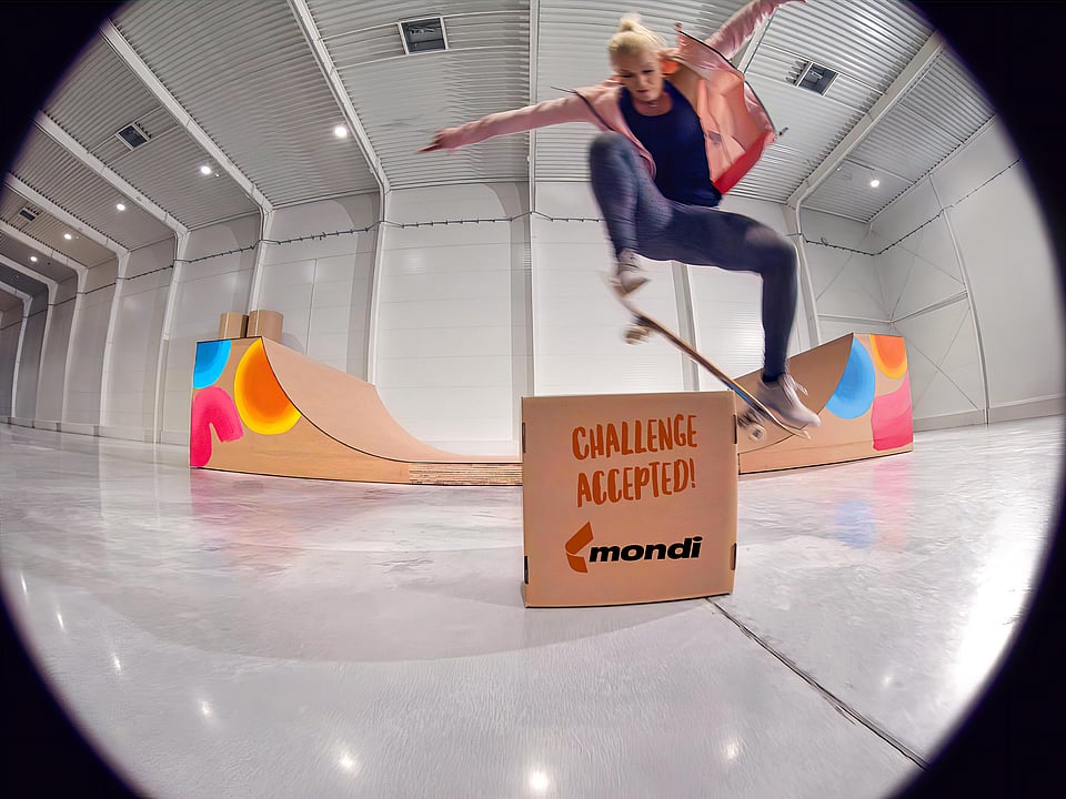 Olympic skateboarder Julia Brückler performing a skateboard trick over a carboard box with "challenge accepted" written on it and designed by Mondi