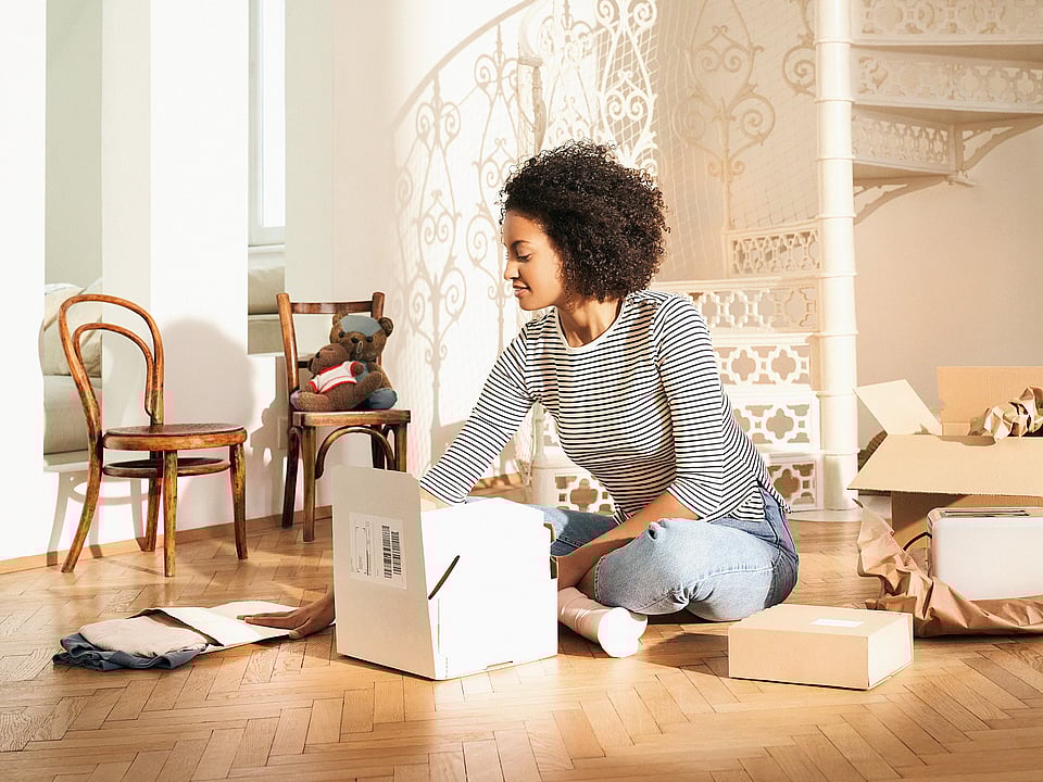 A woman sitting on the floor opening a corrugated box. Other corrugated boxes surround her, as well as a paper mailer bag and filling material made of paper.