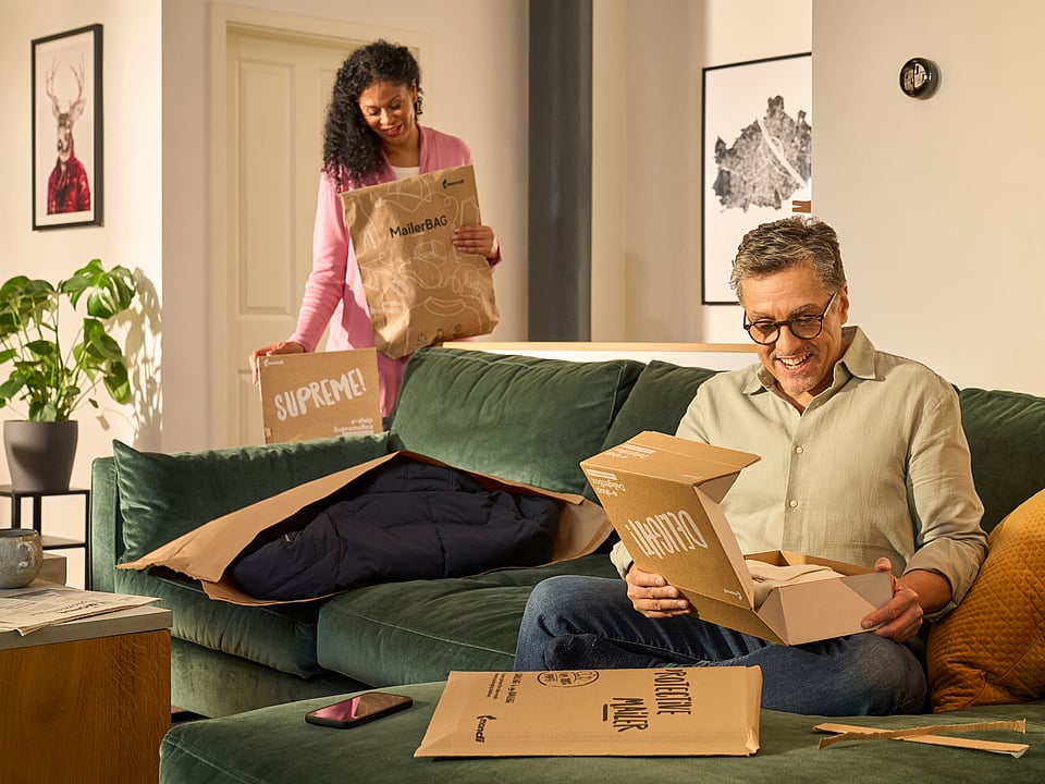 A man is sitting on the sofa unpacking a Mondi DelightBox with a Protective Mailer in front of him, while a woman stands in the back holding a MailerBag and a SupremeBox.