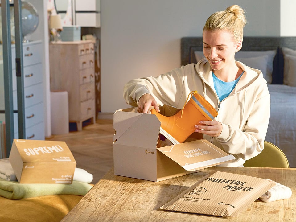 A smiling woman is sitting at a desk, placing a boot into a corrugated box. Other corrugated and paper solutions are laid out around her.